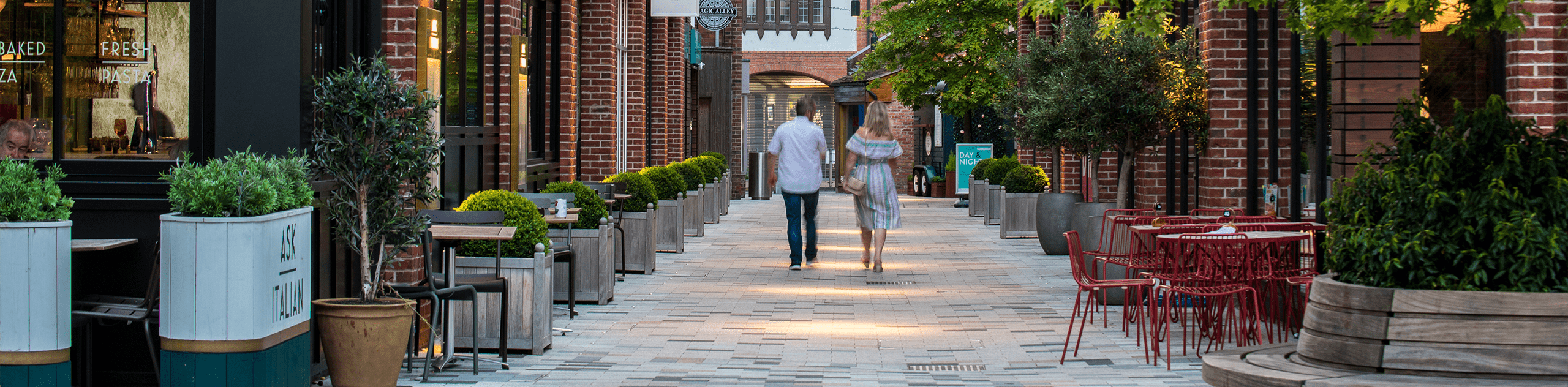 Photograph of couple walking through bell court, featuring resturant spill-out seating and trees in planters. © BMD