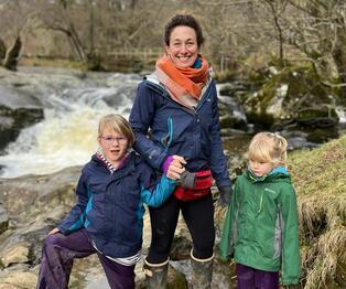 A photograph of Laura Bradley and her two daughters, standing next to a waterfall within a woodland