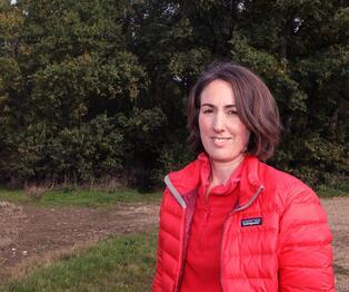 A photograph of a BMD employee wearing a red coat, standing in a field with a woodland backdrop