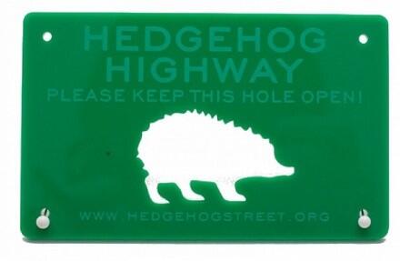 A Green sign with a white hedgehog silhouette on it, the text on the sign says hedgehog highway, please keep this hole open