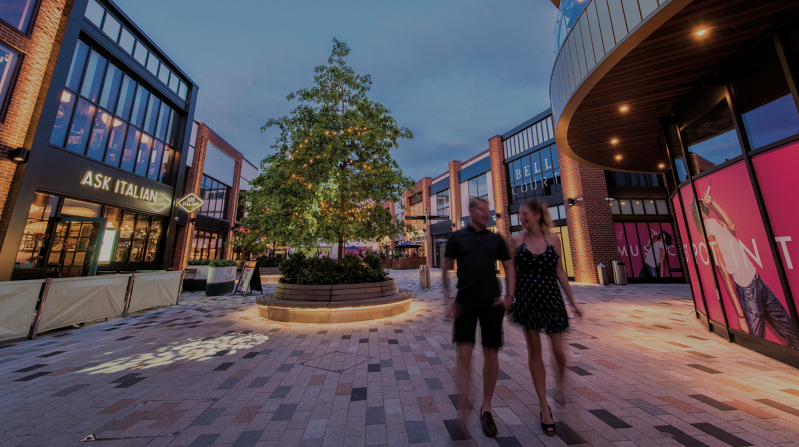 A photograph of a commercial shopping plaza with a mix of restaurants and shops on either side. A couple is walking through the plaza in front of a decorative tree with fairy lights.