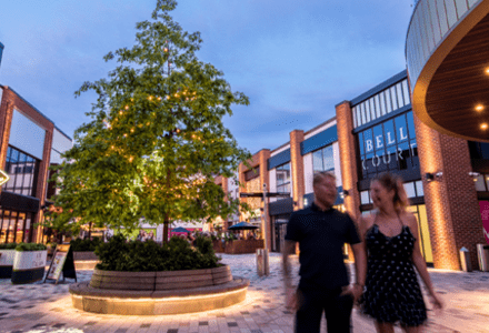 A photograph of a commercial shopping plaza with a mix of restaurants and shops on either side. A couple is walking through the plaza in front of a decorative tree with fairy lights.