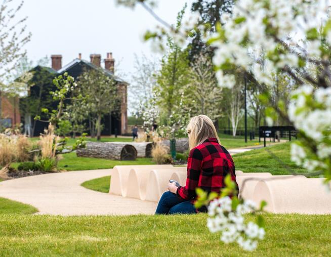 A photograph of a woman sat within an informal green space with play elements and tree planting