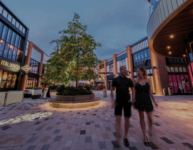 A photograph of a commercial shopping plaza with a mix of restaurants and shops on either side. A couple is walking through the plaza in front of a decorative tree with fairy lights.